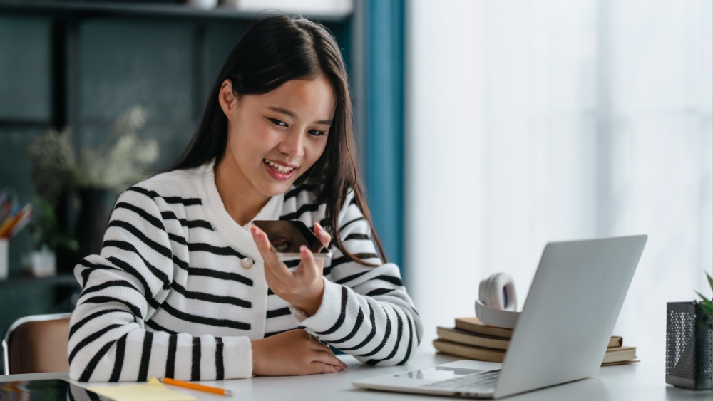 A young woman sitting with a laptop in front of her, talking to the smartphone, smiling.