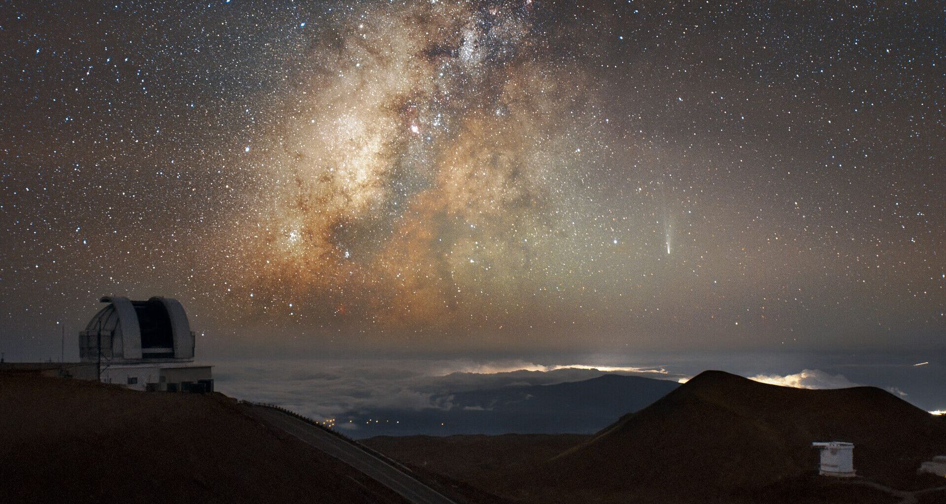 The Milky Way glows orange and yellow amidst the stars in the night sky, with a streak of white at the right side of the image where comet Lemmon moves across with a domed observatory and mountains silhouetted at the bottom