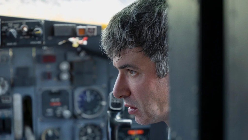 Nathan Fielder sits in the cockpit of an aircraft, staring ahead with intense focus. The background shows various airplane instruments and controls, emphasizing the setting. His expression is serious and contemplative, suggesting a high-stakes or emotionally charged moment.