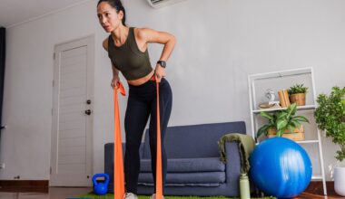 woman in a living room with an orange resistance band under her feet pulling the ends towards her in a bent-over row. there's a blue sofa and big blue balance ball next to her.