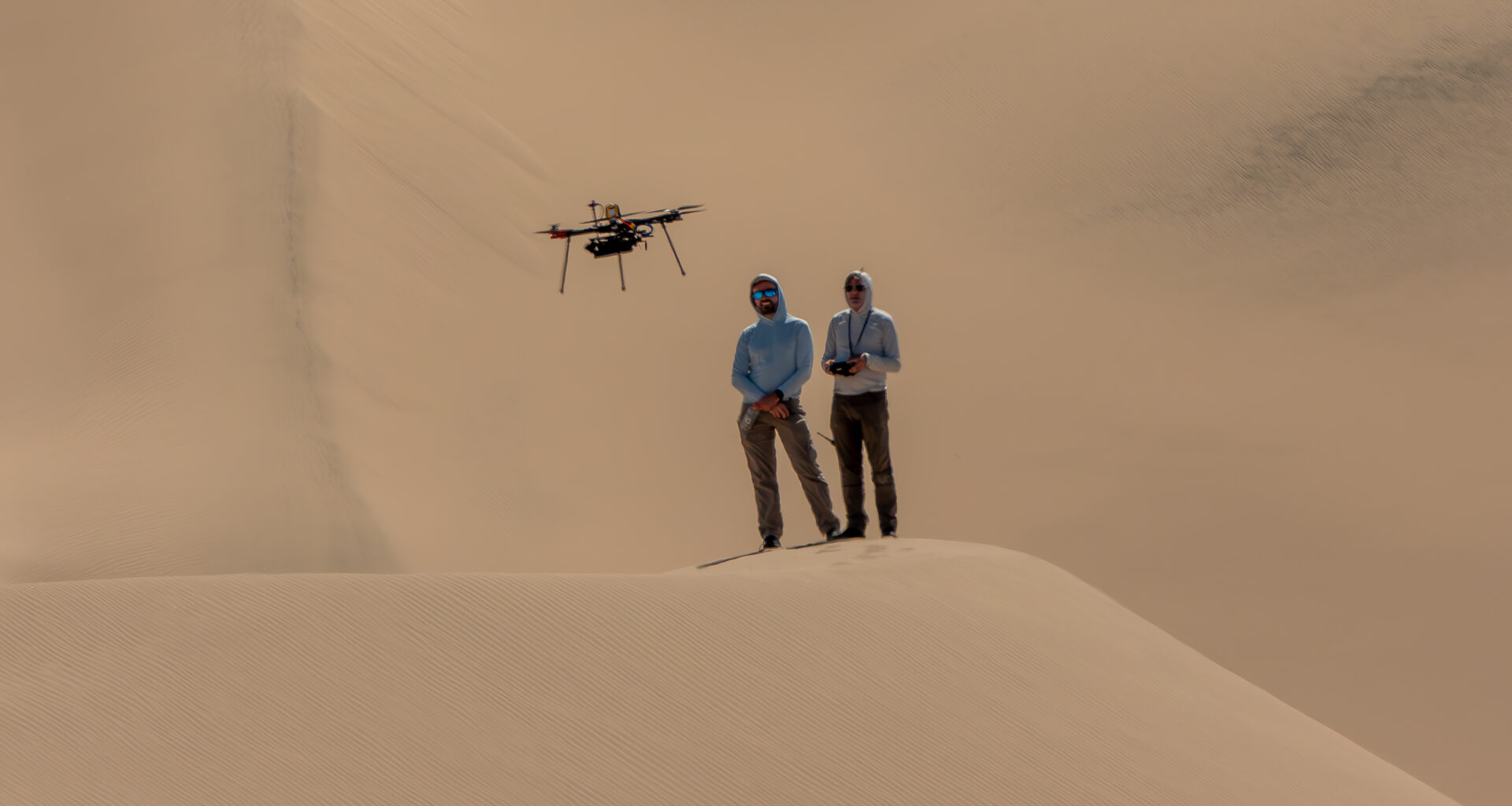Two people stand atop a sand dune, facing the camera. They are both looking at a drone flying in front of them. The person on the right holds a controller. Other sand dunes behind them make up the background of the entire image.