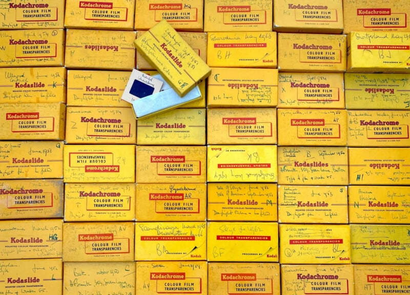 Stacks of yellow Kodachrome film slide boxes, many labeled with handwriting. One box is open, revealing a blue photographic slide inside. The boxes fill the entire image, creating a colorful, vintage pattern.
