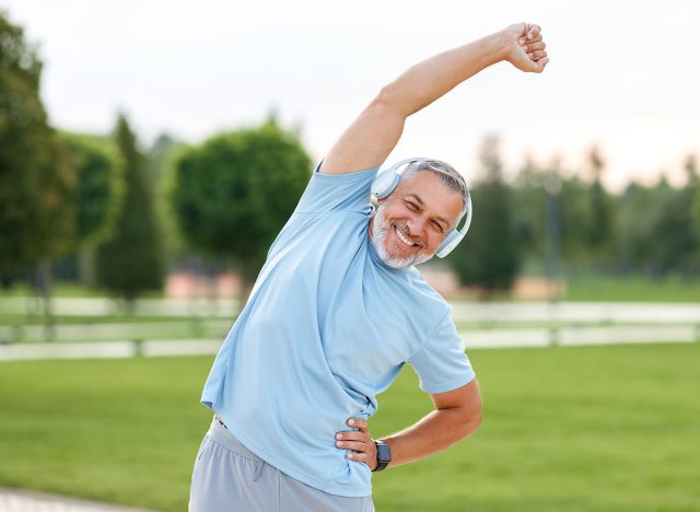Staying active after retirement. Happy joyful mature retired sportsman wearing headphones and sportswear doing side stretching exercises with arm over his head, exercising outside in city park