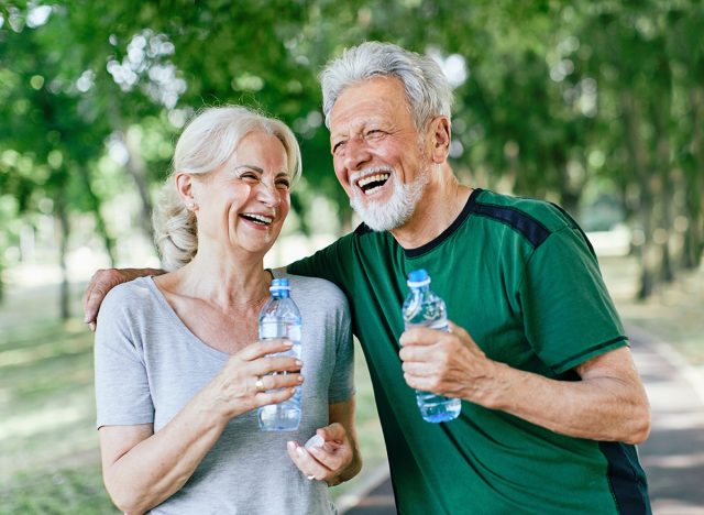 Smiling active senior couple holding water bottles, drinking and jogging together in the park