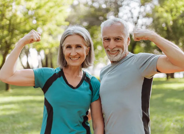 Smiling, attractive senior athletic couple flexing muscles in park, looking at camera outdoors, active lifestyle concept