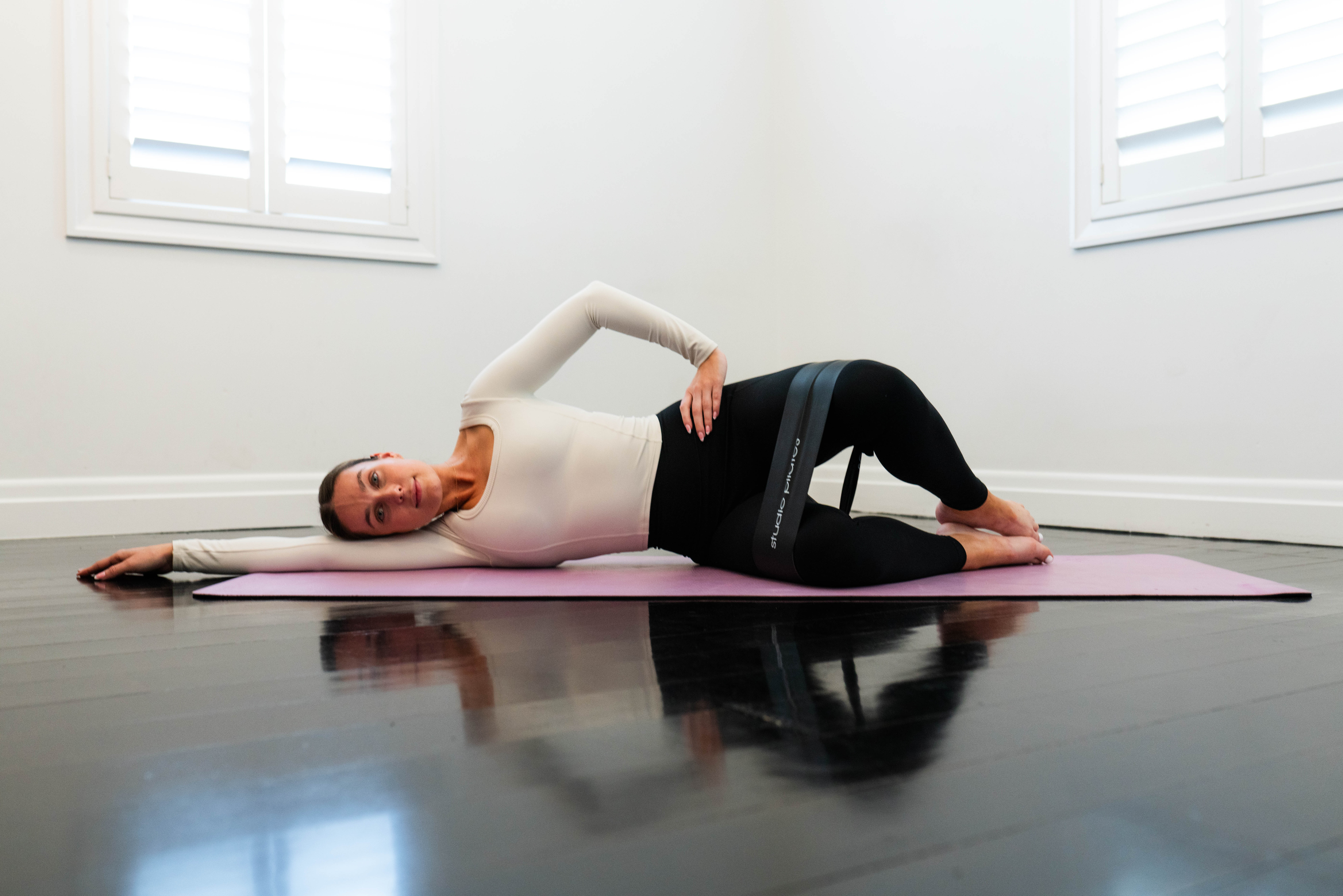 A model from Studio Pilates performs a banded clamshell on an exercise mat. She is lying on her right side, left arm extended on the floor above her head, knees bent and feet together with a band around her thighs. Her left knee is raised above her hip.
