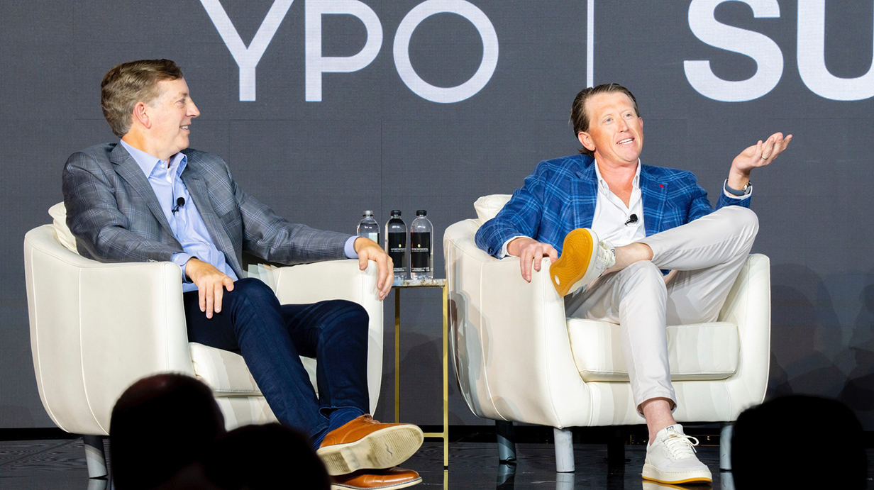A man seated on a stage gestures as he talks as another man seated nearby watches.