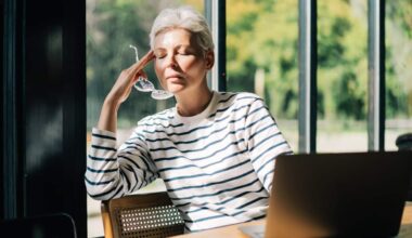 Woman with eyes closed touches her hand to her temple, holding her glasses in the same hand, as she sits in a sunny spot inside at a table with her laptop.