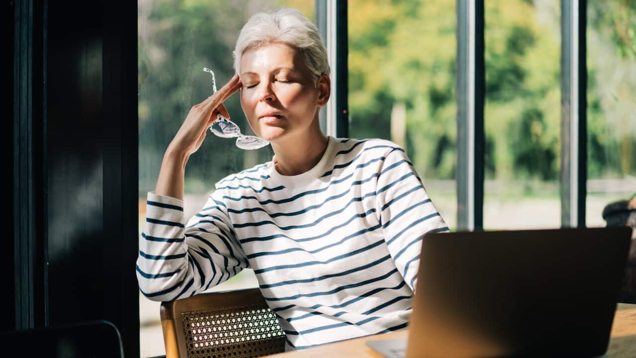 Woman with eyes closed touches her hand to her temple, holding her glasses in the same hand, as she sits in a sunny spot inside at a table with her laptop.