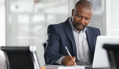 An investor scribbling notes into a notebook while seated at a desk in front of a computer.