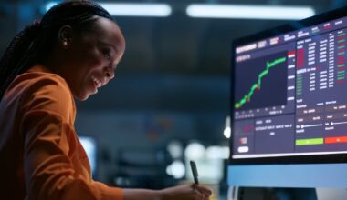 A smiling person writing notes while looking at stock charts on the computer.