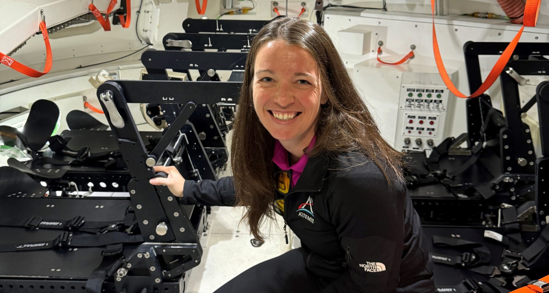 A woman crouches inside the mockup of a spacecraft that is equipped with seats for crewmembers.