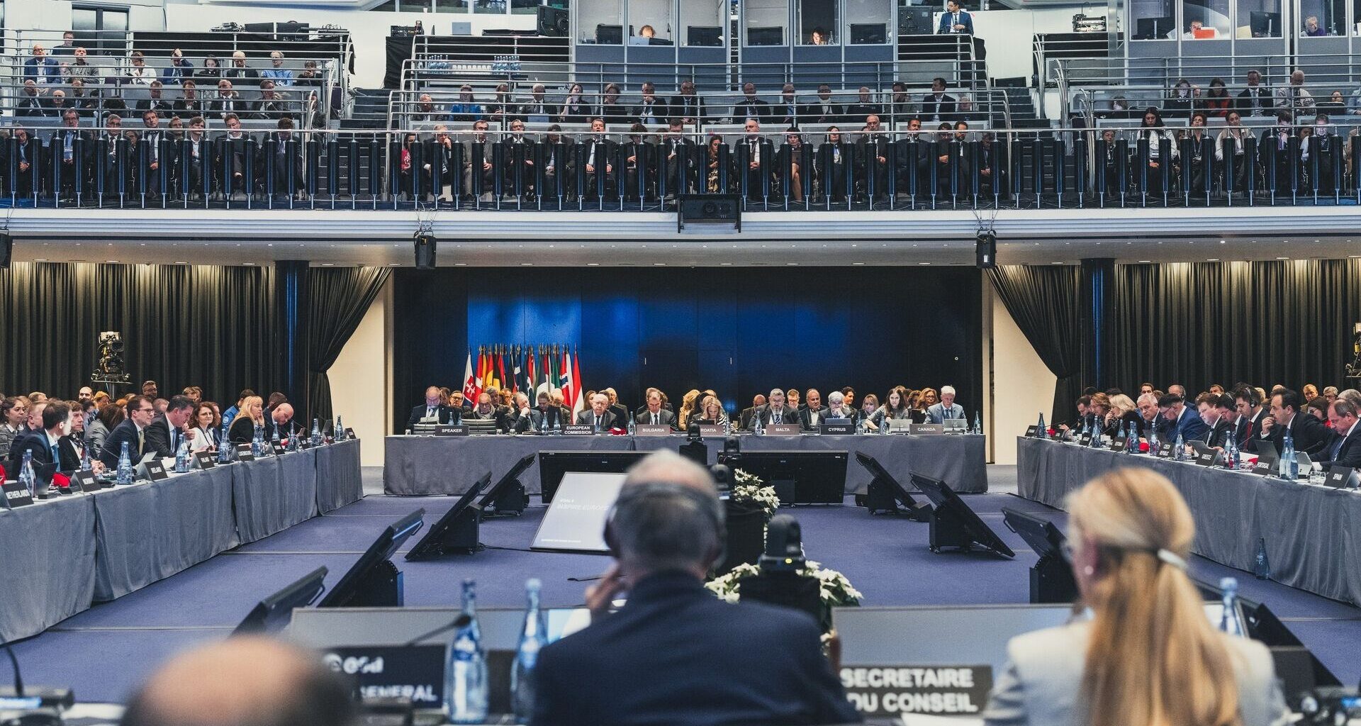 A series of people sit at four different tables making a square to face each other with various flags of their corresponding countries next to them.
