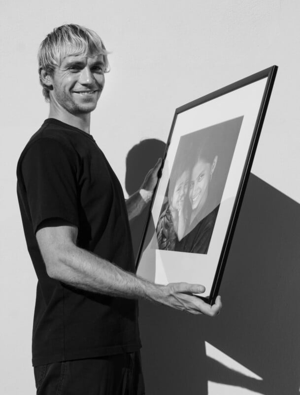 A smiling man with light hair holds a framed black-and-white photo of two people, standing against a plain wall in bright sunlight.