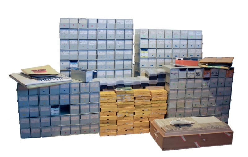 Stacks of archive boxes and folders arranged in rows, with labels and colored dots for identification. Some boxes are open, and packets and envelopes are scattered on top. The background is plain white.