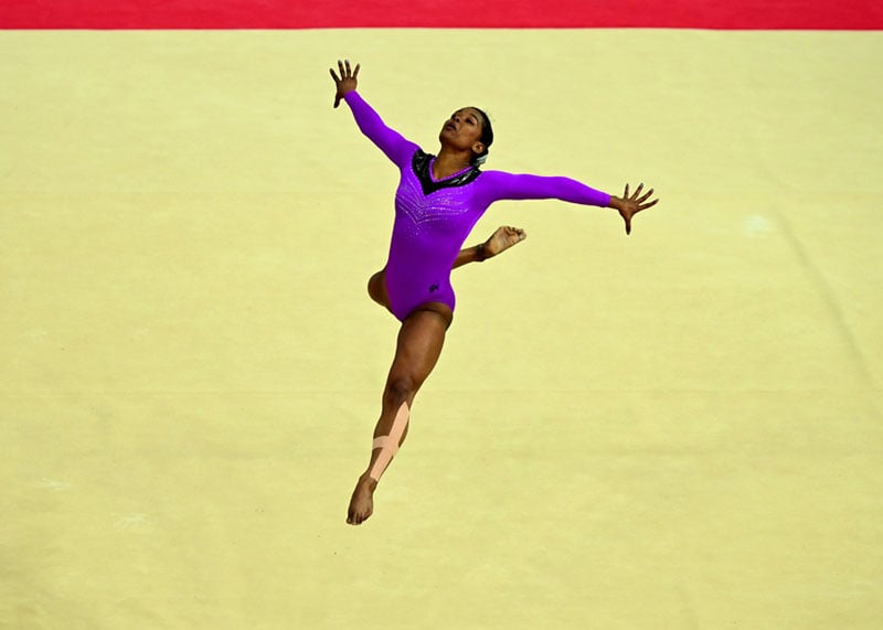 A gymnast in a purple leotard performs a leap on a beige floor, arms extended and one leg bent behind her, with a red border visible at the top of the image.