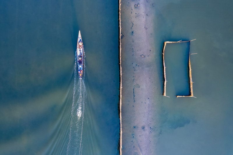 Aerial view of a boat moving through blue-green water, leaving a wake behind, with a narrow barrier and a rectangular enclosure in the water to the right.