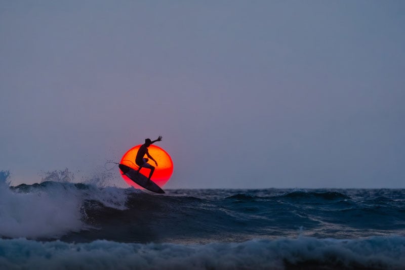 A surfer rides a wave at sunset, silhouetted against the orange sun low on the horizon, with dark waves and a clear sky in the background.