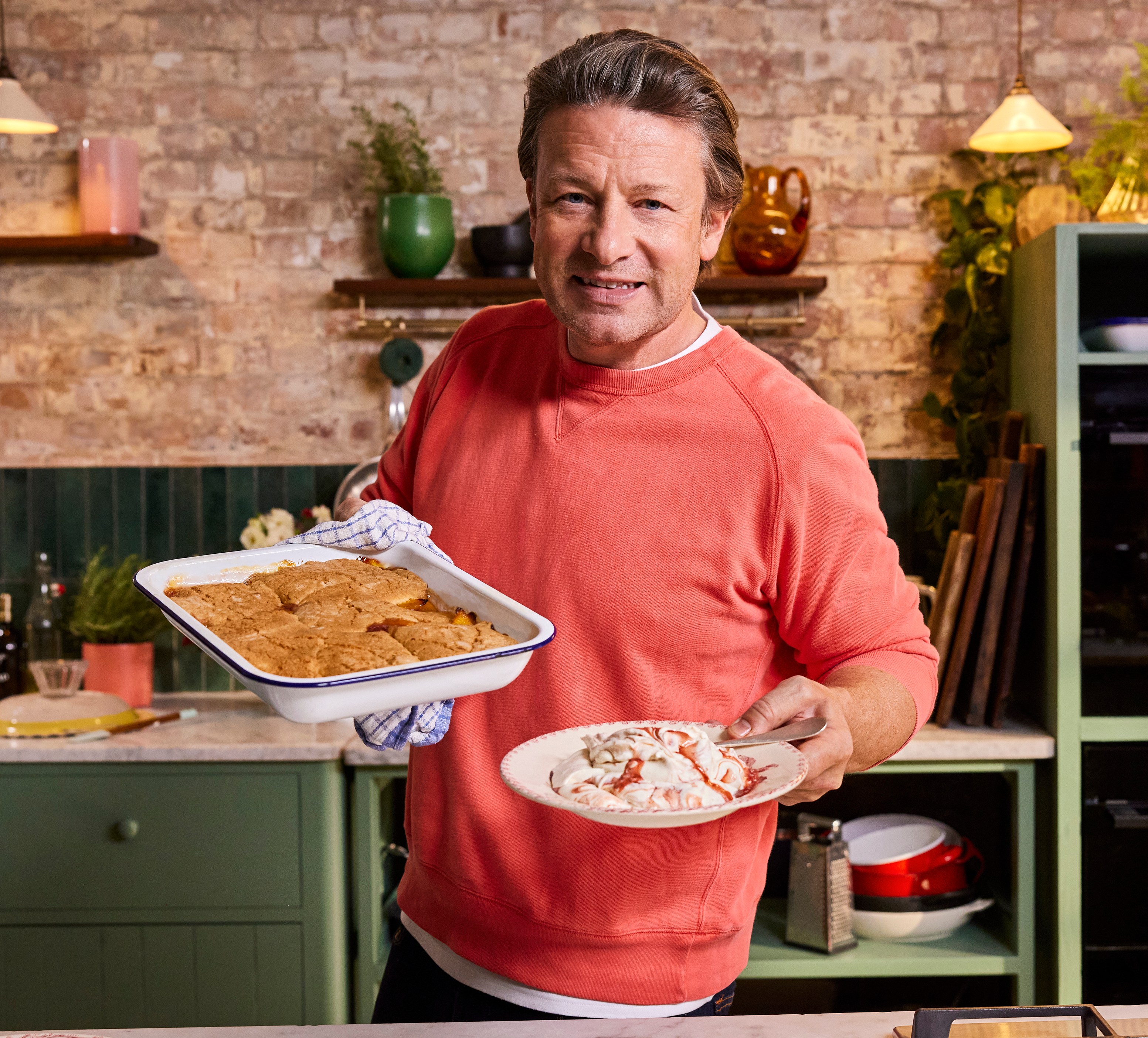 Jamie Oliver holding a baking dish of cobbler and a plate with whipped cream and syrup.