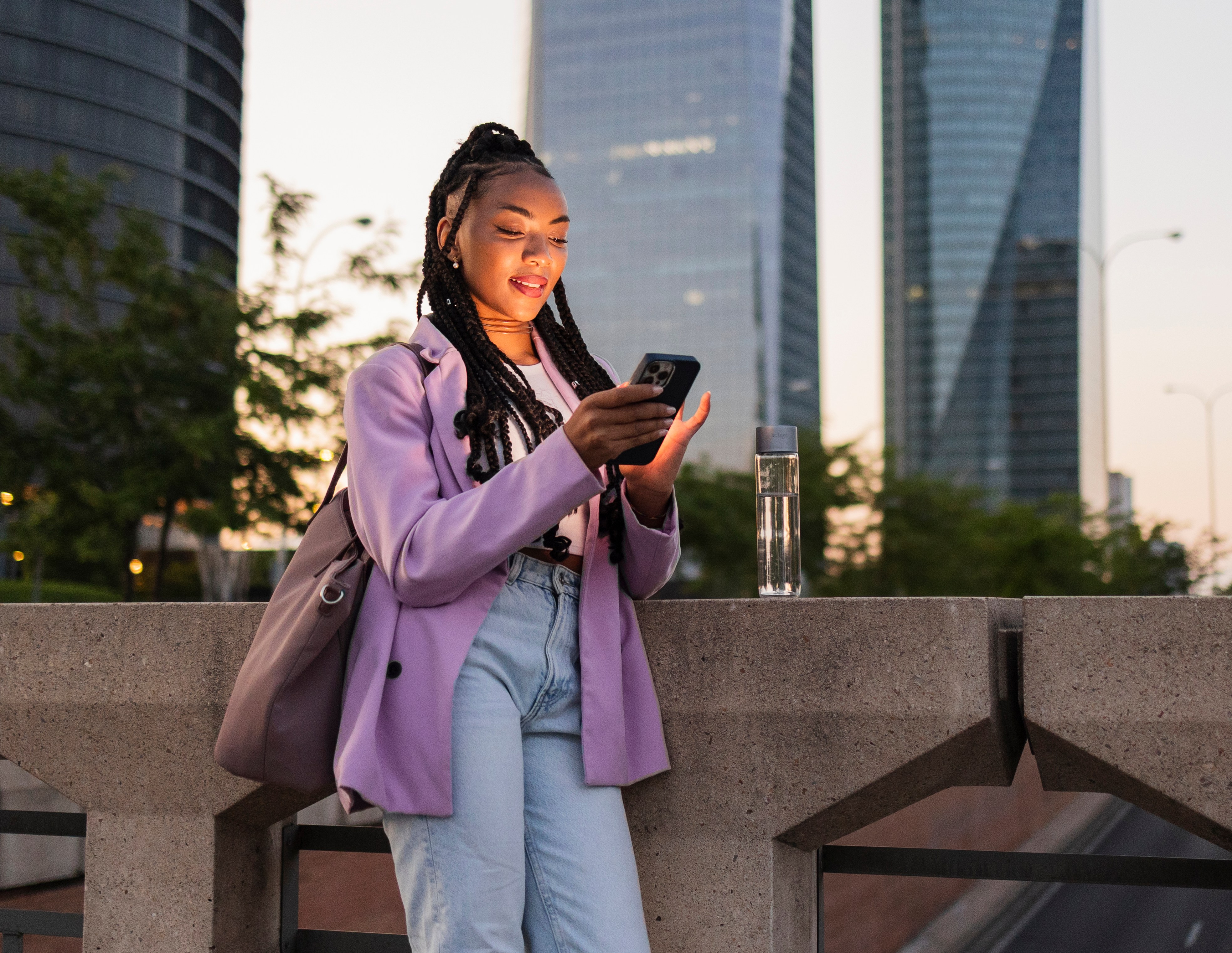 Young businesswoman smiling and using her smartphone on a bridge railing with skyscrapers in the background at sunset.