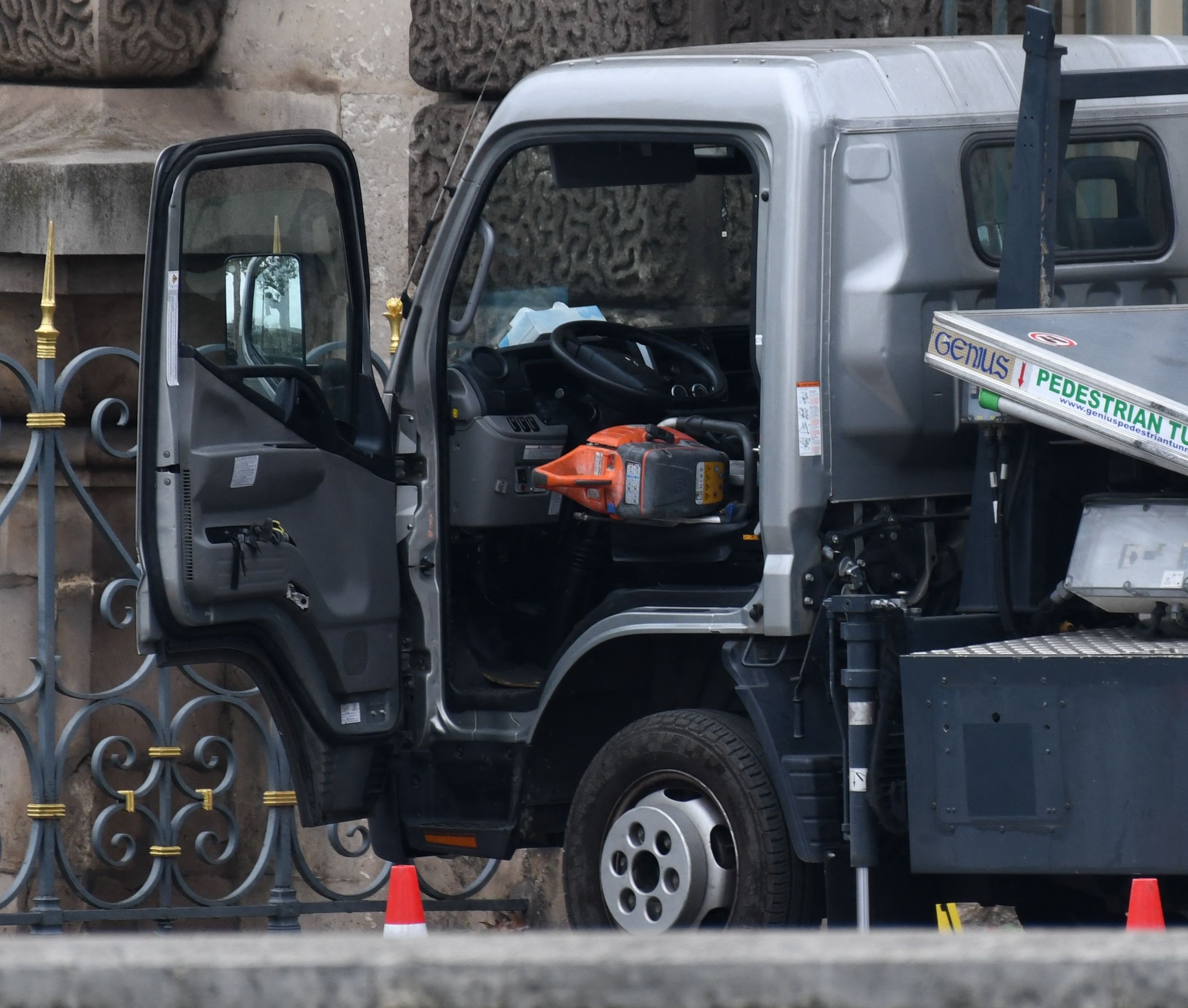 A gray truck with an open door is parked beside a decorative metal fence and stone building. An orange chainsaw is visible in the driver's seat.