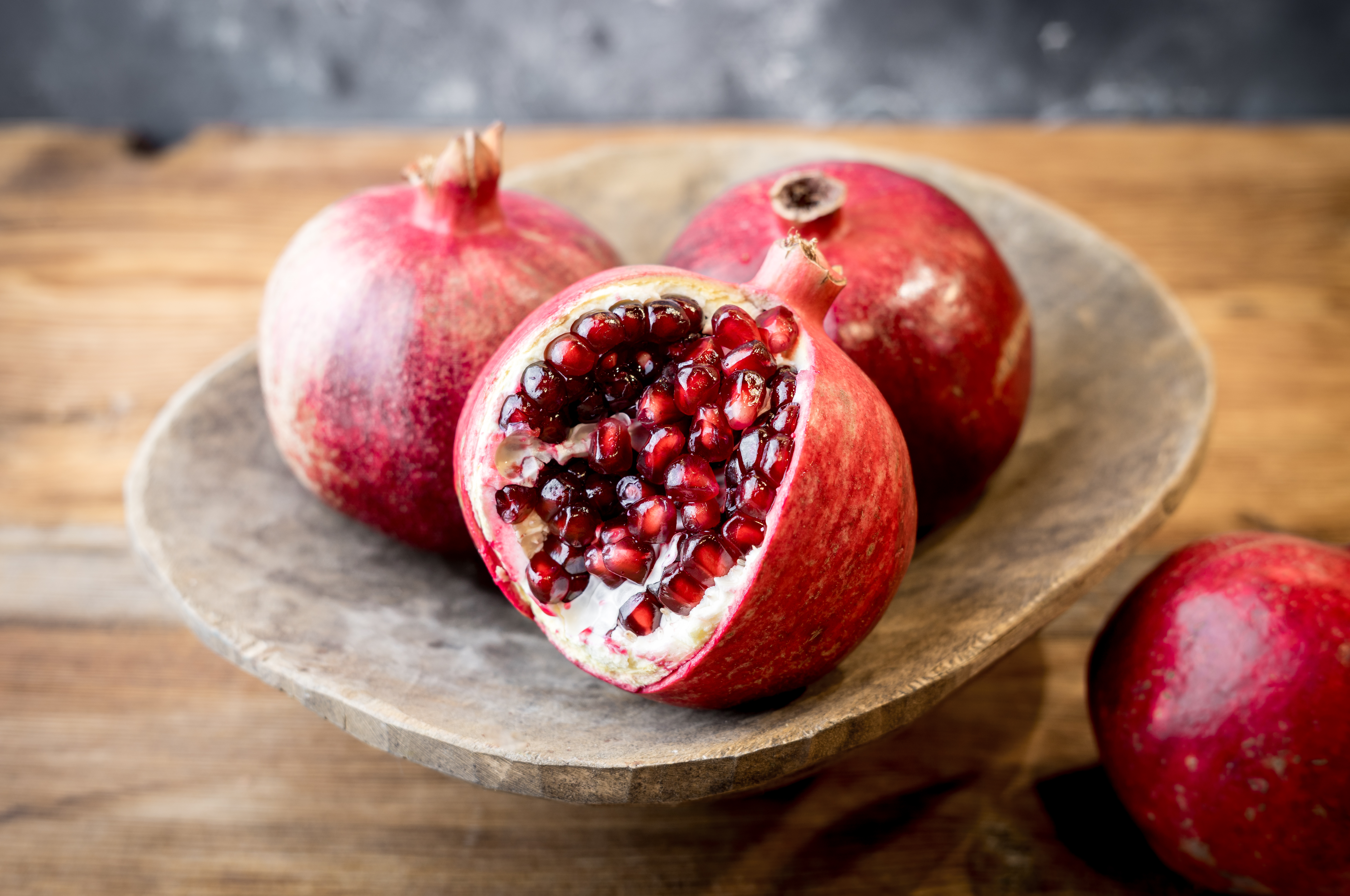 Close-up of three pomegranates on a wooden table, one of which is cut in half revealing its seeds.