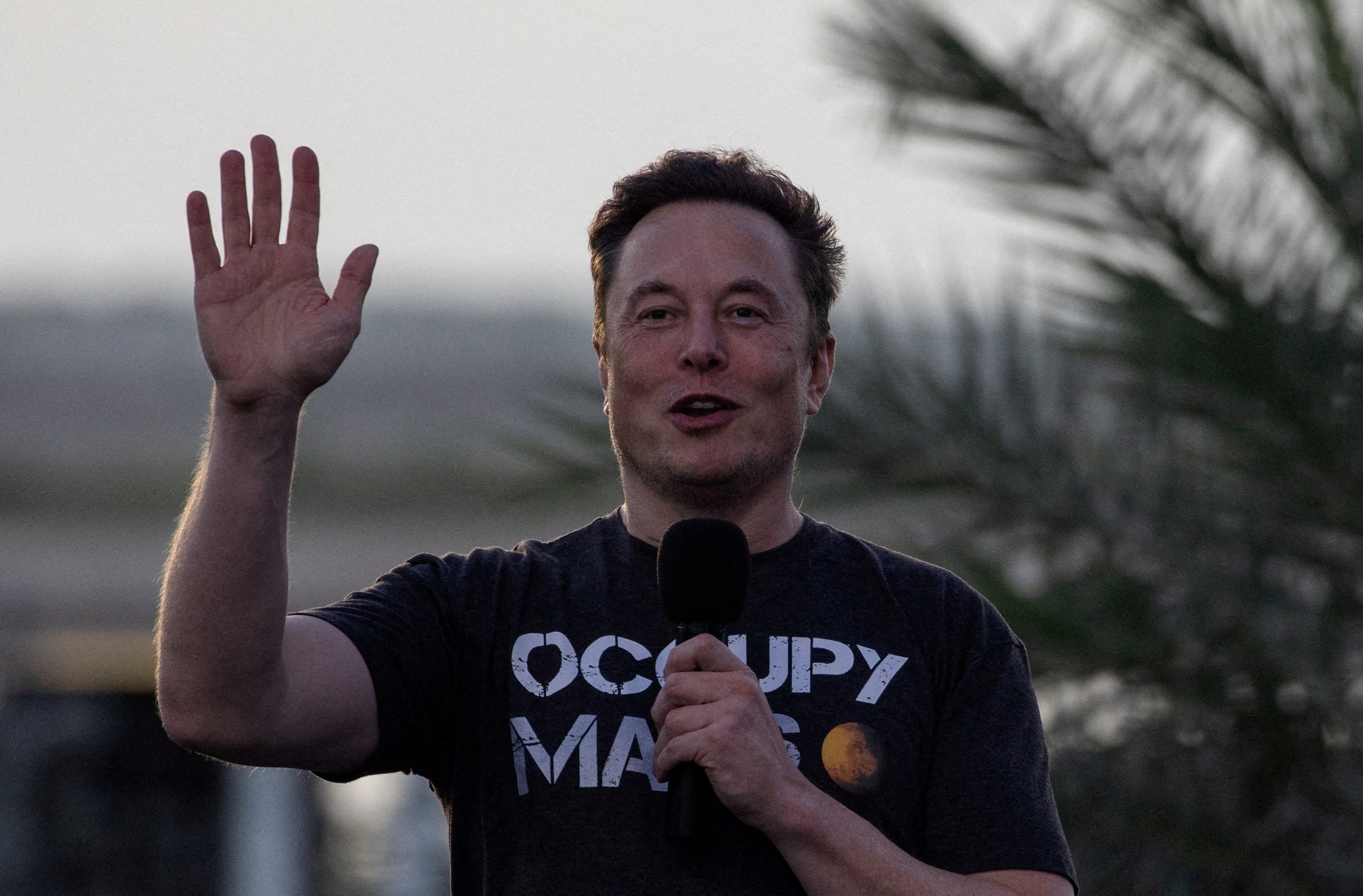 Elon Musk speaking into a microphone and gesturing with his right hand.