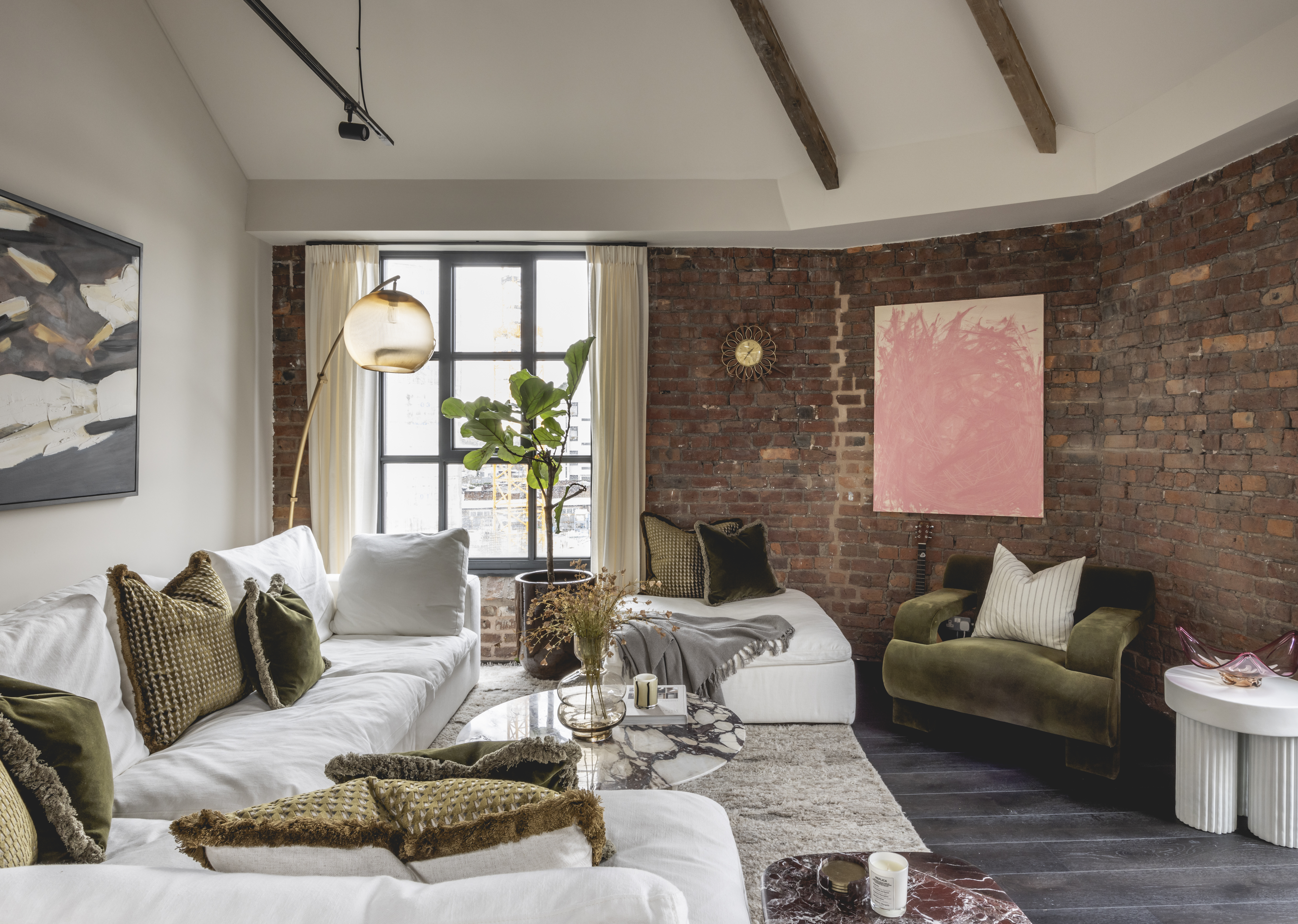 Victoria Magrath's apartment living room designed by Jolie Studio, featuring white couches, velvet pillows, exposed brick walls, and a large window.