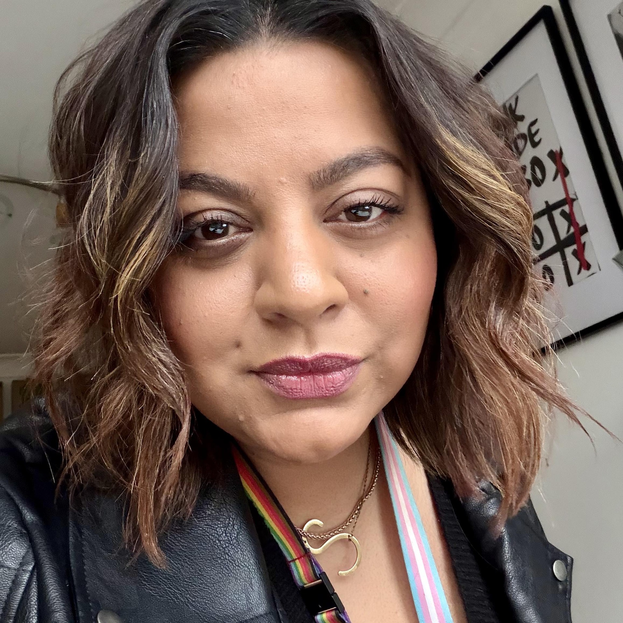 Saima Akhtar wearing a black leather jacket, with rainbow-striped and blue-pink-white striped lanyards, and a gold 'S' necklace.