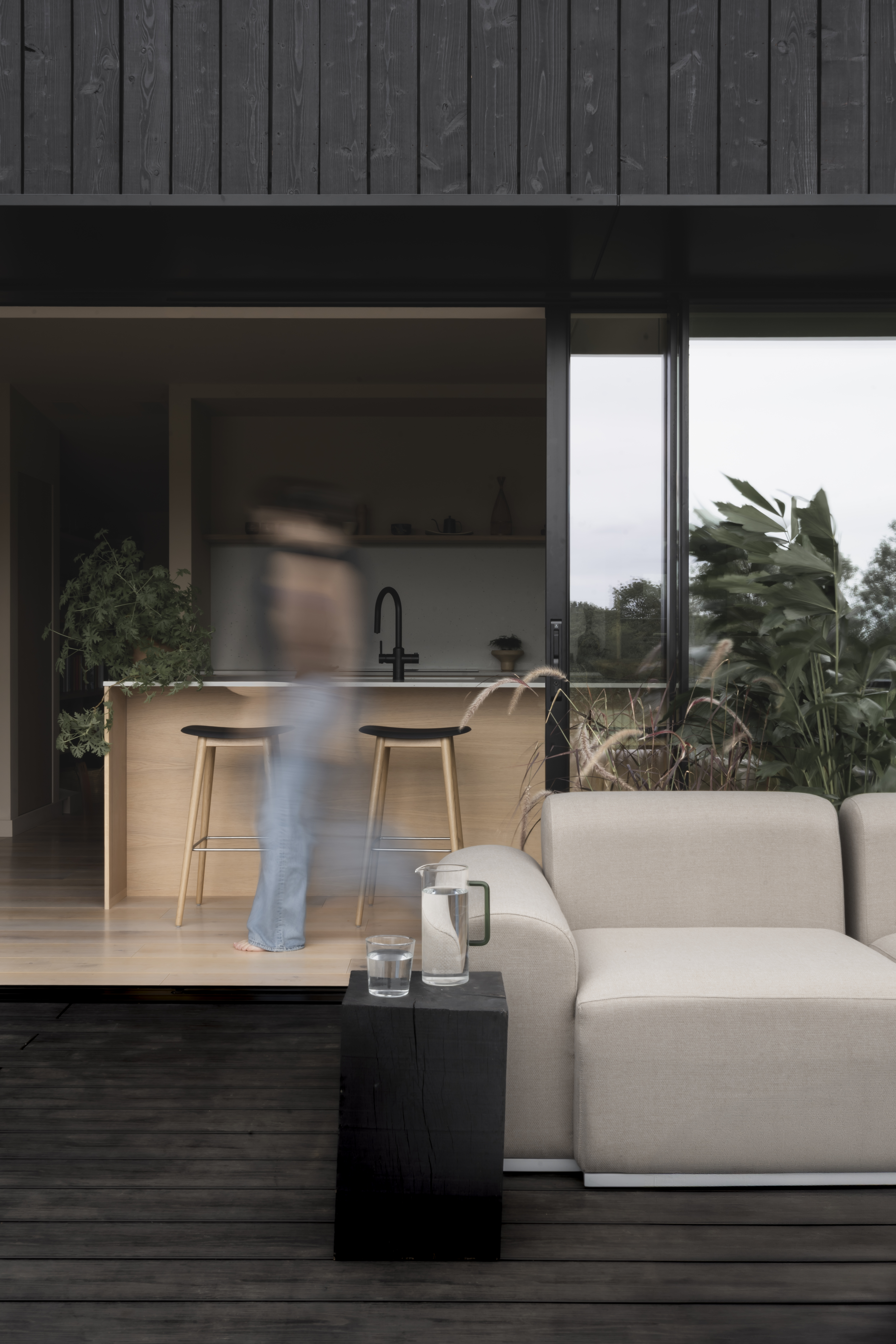 A person walks barefoot in a modern open-plan home from a kitchen with a wooden island and stools to a living area with a cream sofa and black wooden block table, surrounded by lush green foliage outside.