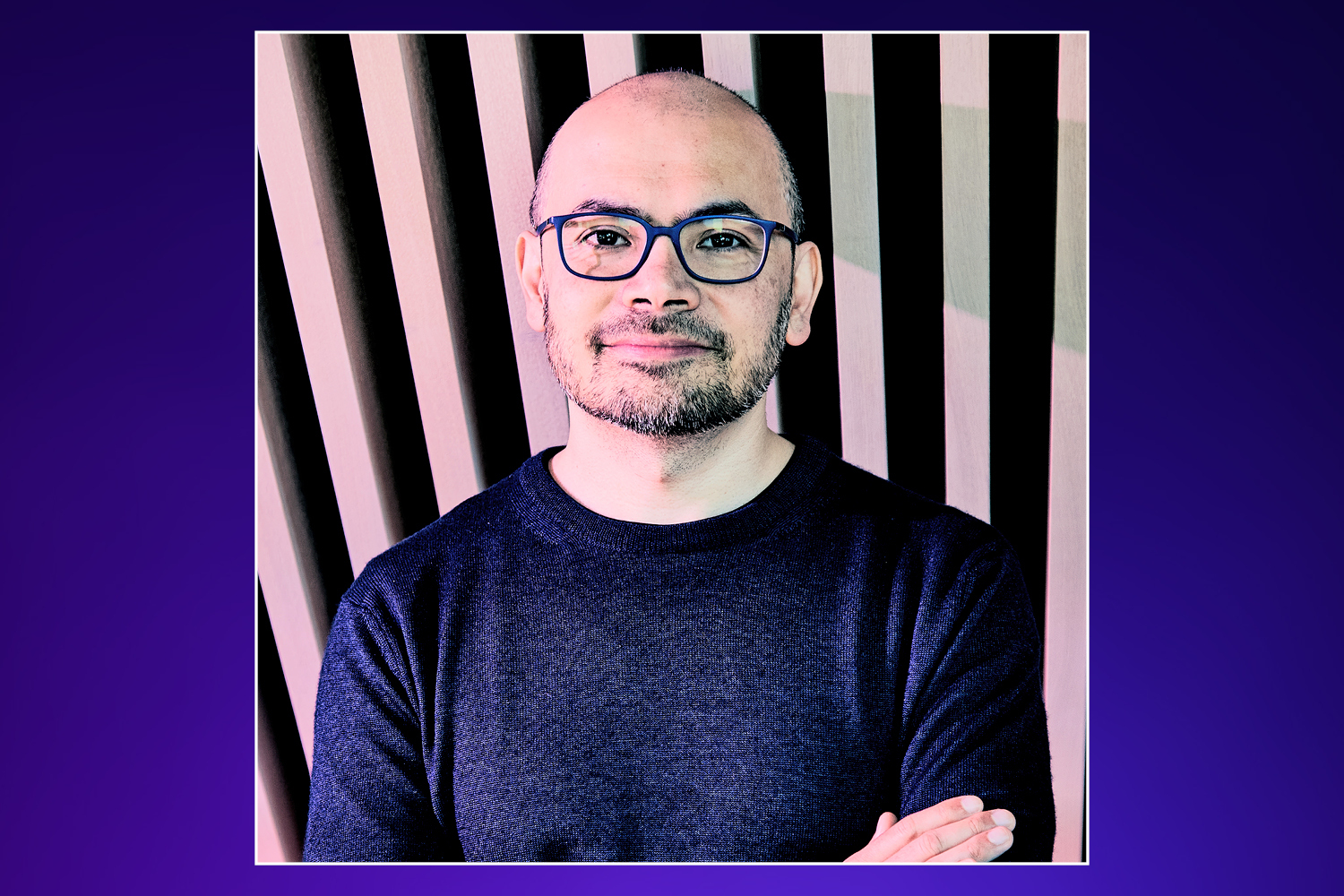 Man with glasses and a beard in a dark sweater, arms crossed, standing against a striped background.