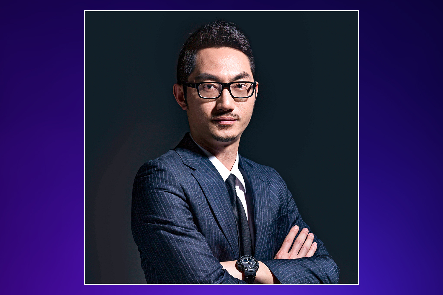 A man in a pinstripe suit, white shirt, and black tie with glasses and a small mustache looking at the camera with his arms crossed.