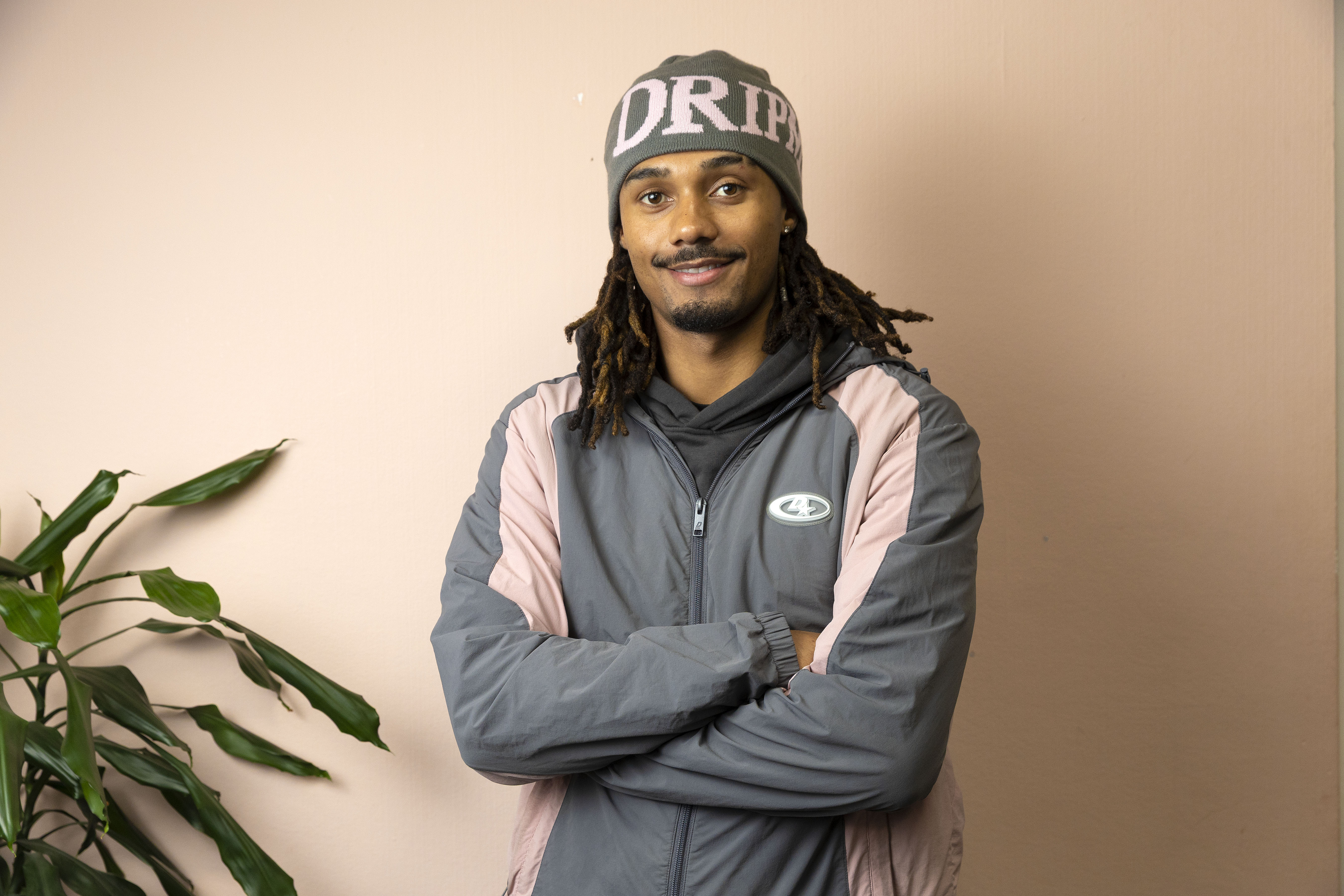 Ash in a gray and pink jacket and a gray beanie with the word "DRIP" on it, smiling.