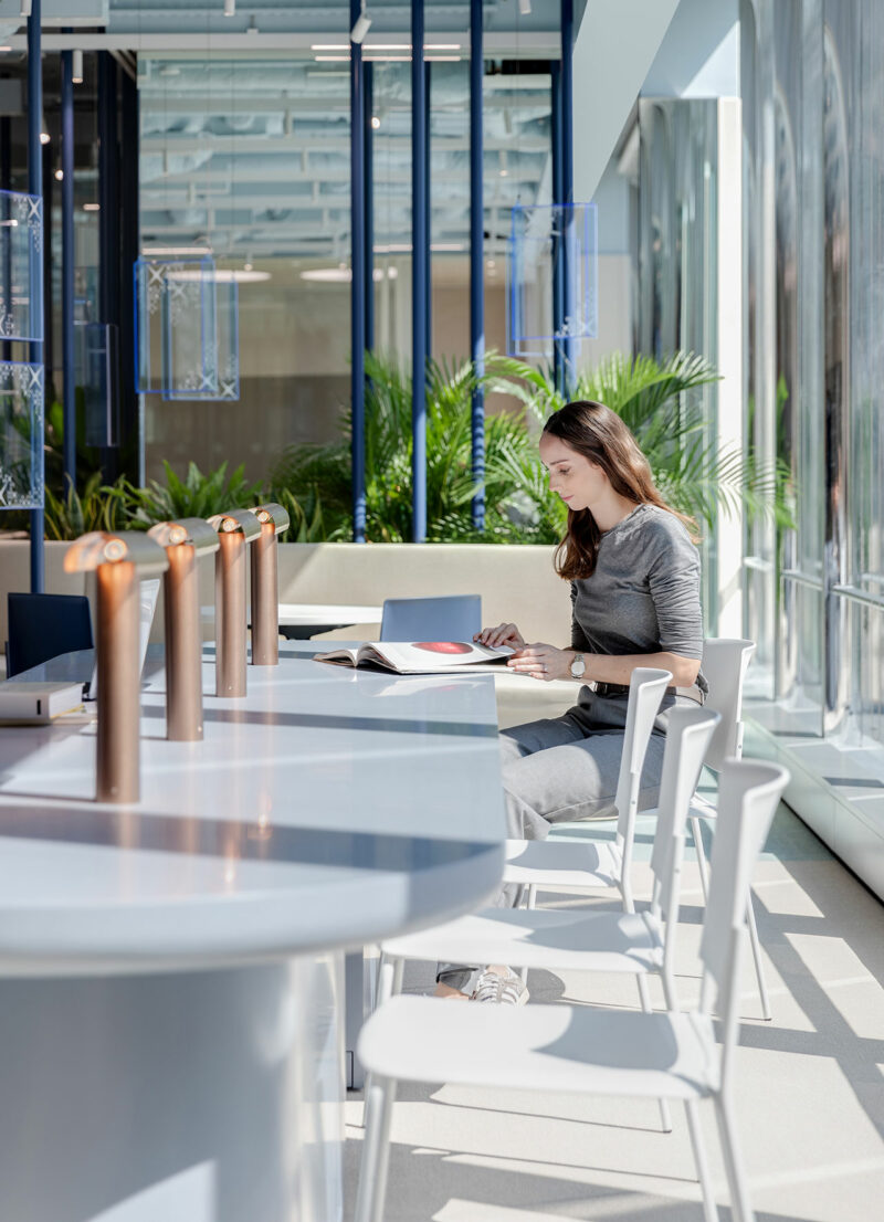 A woman sits alone at a modern table in a bright, glass-walled room, reading a book with greenery visible outside.