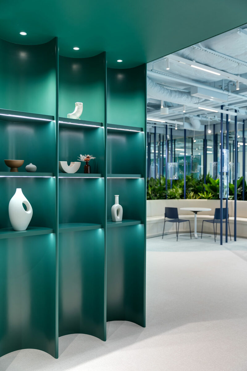 A modern office space with a green curved shelving unit displaying minimalist vases and bowls, adjacent to a seating area with blue chairs and indoor plants.