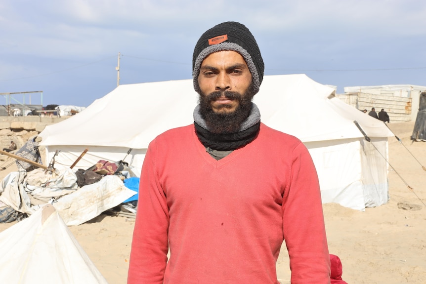 A bearded man wearing a hat and scarf standing in front of tents pitched on sand.