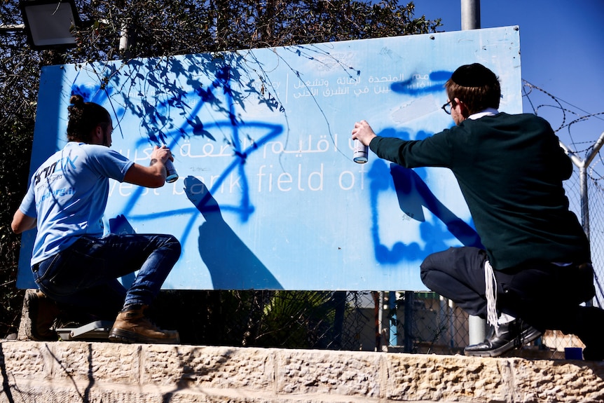 Two men spray paint over a UNRWA sign.