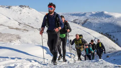 David Murch/Montane Spine Race Endurance racers with walking poles make their way up a steep slope in the snow
