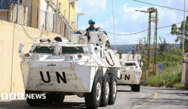 UN peacekeeping force in Lebanon. Two large white vehicles drive around a winding road. A UN officer in bright blue helmets can be seen stood on each vehicle.