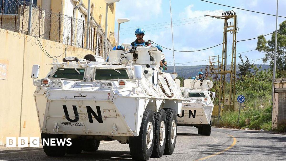 UN peacekeeping force in Lebanon. Two large white vehicles drive around a winding road. A UN officer in bright blue helmets can be seen stood on each vehicle.