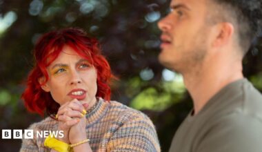 Traitors contestant Jessie Stride is seen chatting to fellow faithful Matthew. Jessie has bright red hair, yellow and orange eyeshadow and is wearing a rainbow jumper. The background has trees.
