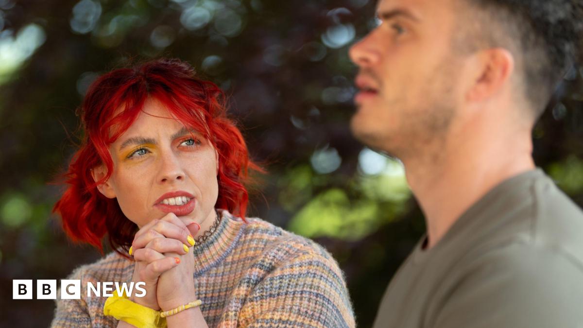 Traitors contestant Jessie Stride is seen chatting to fellow faithful Matthew. Jessie has bright red hair, yellow and orange eyeshadow and is wearing a rainbow jumper. The background has trees.