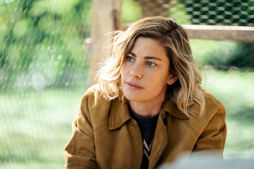 A TV still of Brooke Satchwell, 45, in close-up. She's wearing a coat, looks concerned, and is sitting inside a chicken coop.