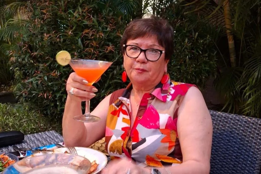 A middle-aged woman raises a cocktail as she sits at an outdoor cane table.