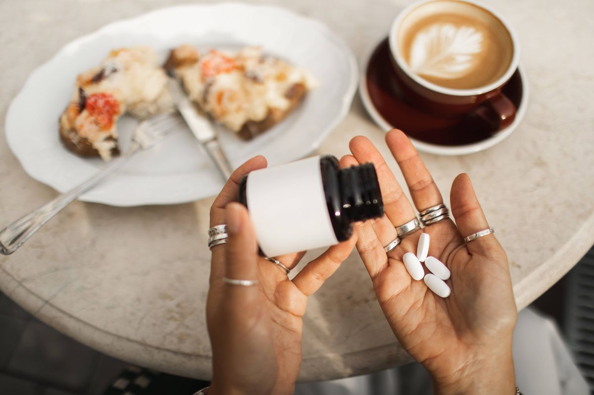 Close-up of female hands holding a pill bottle with pills in the palm, beside a coffee cup and a plate with cake, illustrating a casual morning routine and wellness theme.