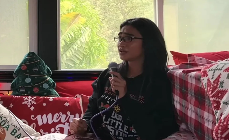 Bretman Rock in a festive room with holiday-themed pillows and a small Christmas tree, holding a microphone, wearing a patterned sweater