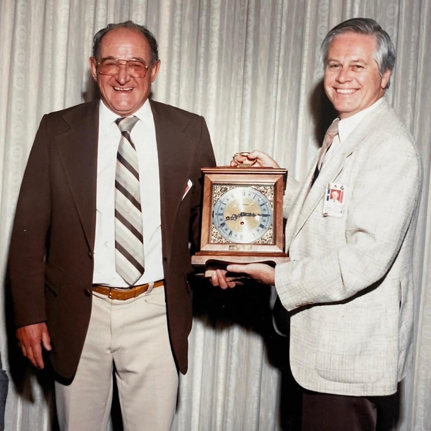 Two middle-aged men stand smiling at the camera wearing jackets and ties and holding an ornate clock in a wooden frame.