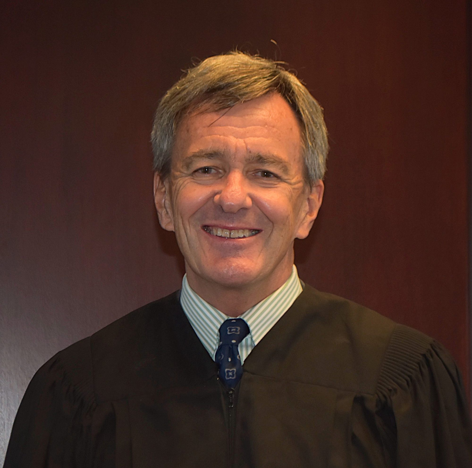 A middle-aged man wearing a black judicial robe and a tie stands smiling in front of a plain dark wooden background.