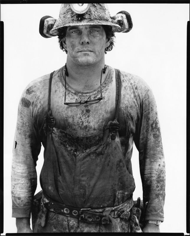 A miner wearing a helmet with a headlamp stands facing the camera. His face, clothes, and gloves are covered in dirt, and he has a serious expression, with safety glasses hanging around his neck.