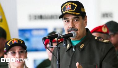 Venezuela’s leader Nicolas Maduro, wearing a military uniform, stands in front of two microphones as he attends a year-end salutation to military forces his wife Cilia Flores.